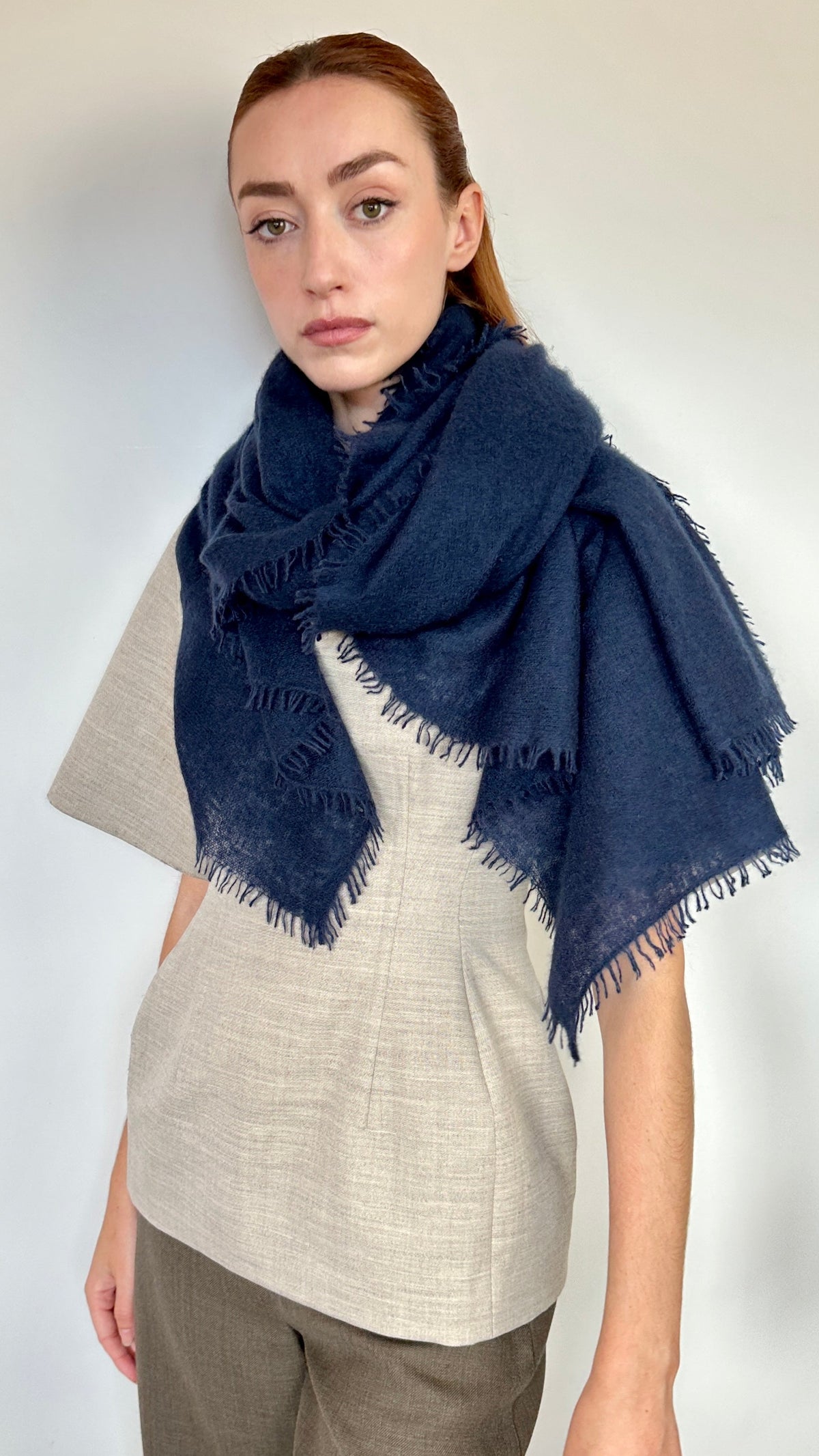 A woman modeling a dark navy blue cashmere fringed scarf wrapped around her shoulders, paired with a light beige linen top and brown trousers, against a plain off-white wall.