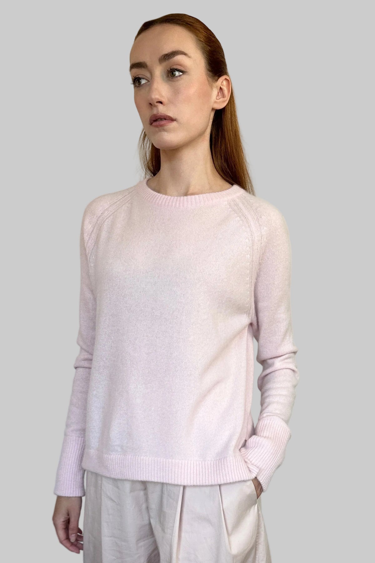 A woman modeling a light pink long-sleeve knit sweater and matching pale blush wide-leg trousers, standing against a plain off-white wall with light wood flooring.