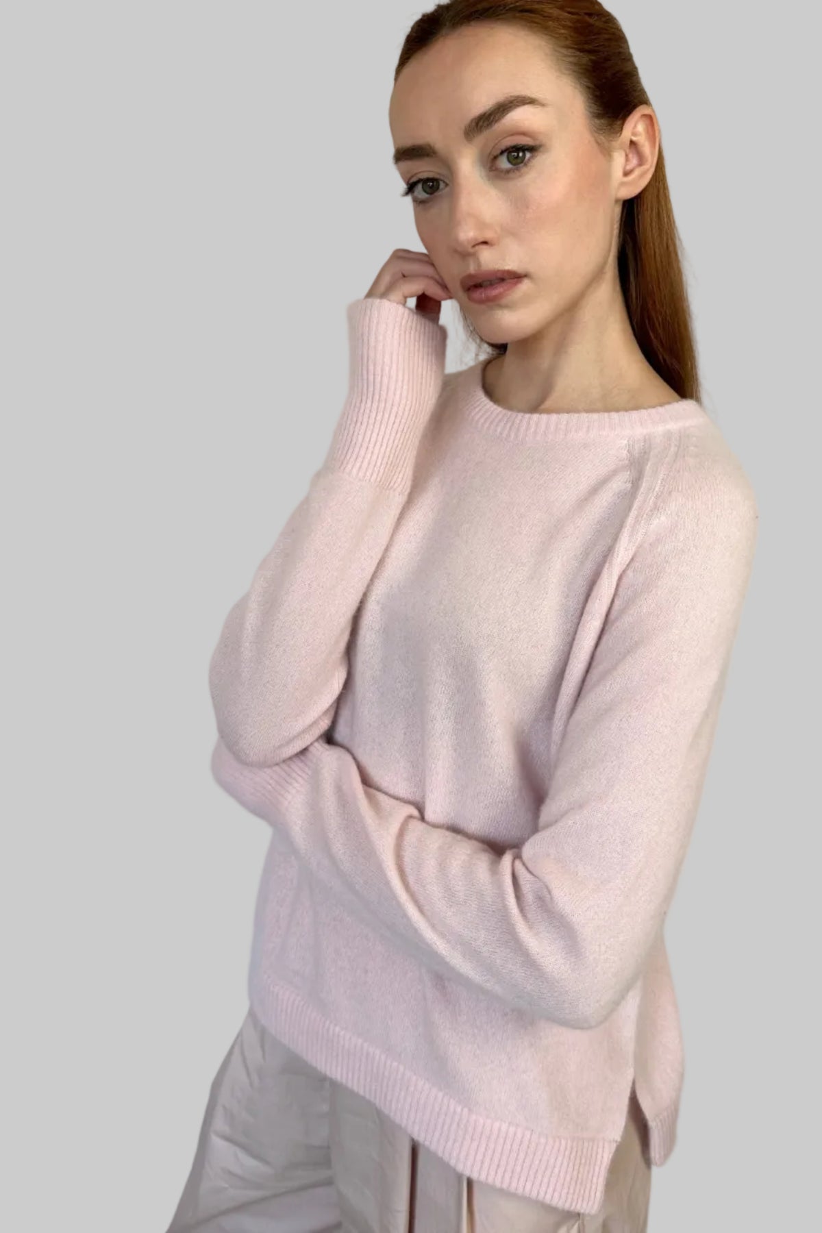 A woman wearing a light pink crewneck knit sweater and pale beige trousers, posing against a plain light blue-gray wall.