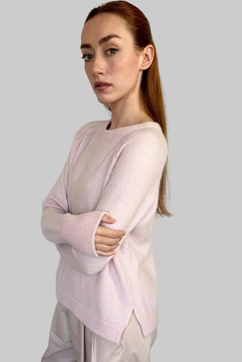 A woman with red hair tied back wears a light pink knit sweater and pale beige trousers, standing against a plain off-white wall with her arms crossed.