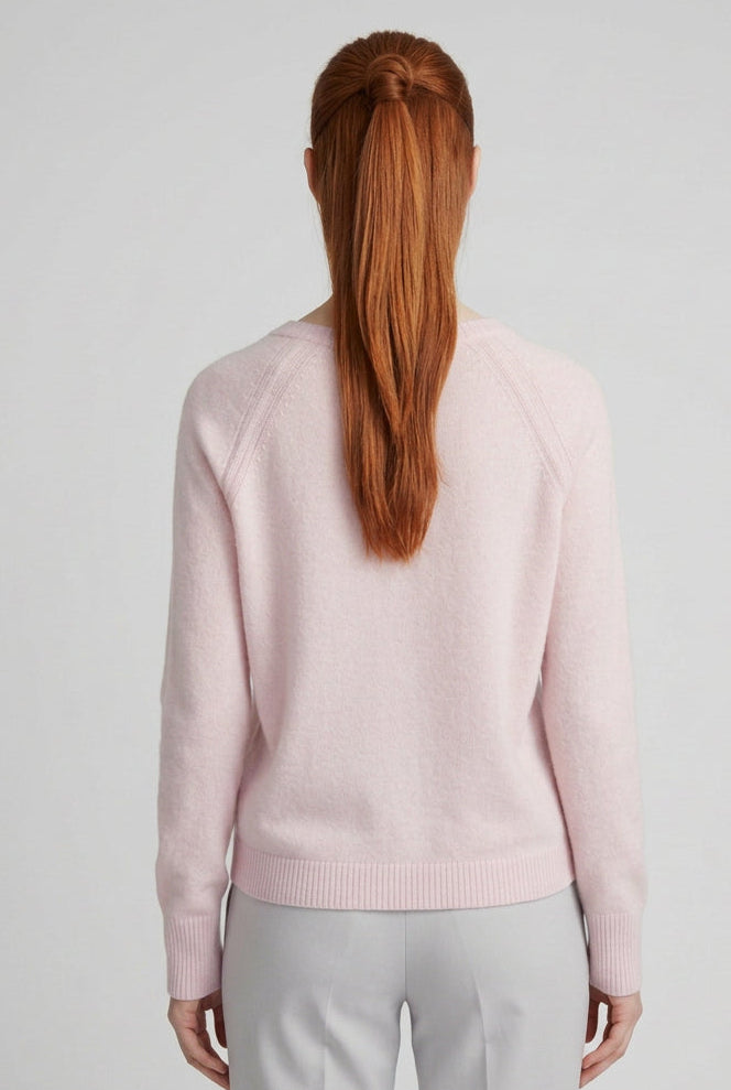 A woman with red hair tied back is shown from behind wearing a light pink knit sweater and beige trousers, standing against a plain off-white wall.