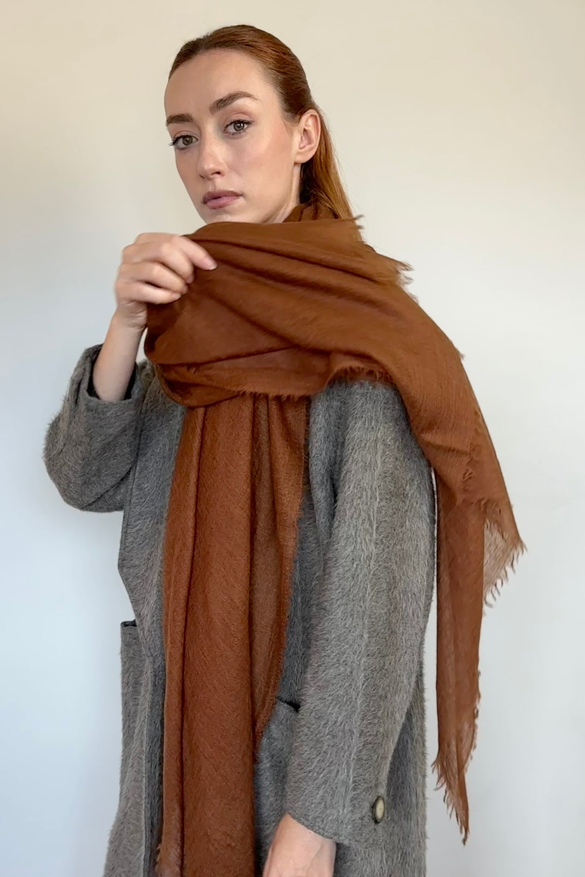 A woman wearing a textured gray coat and modeling a rust-brown fringed scarf against a plain off-white background.