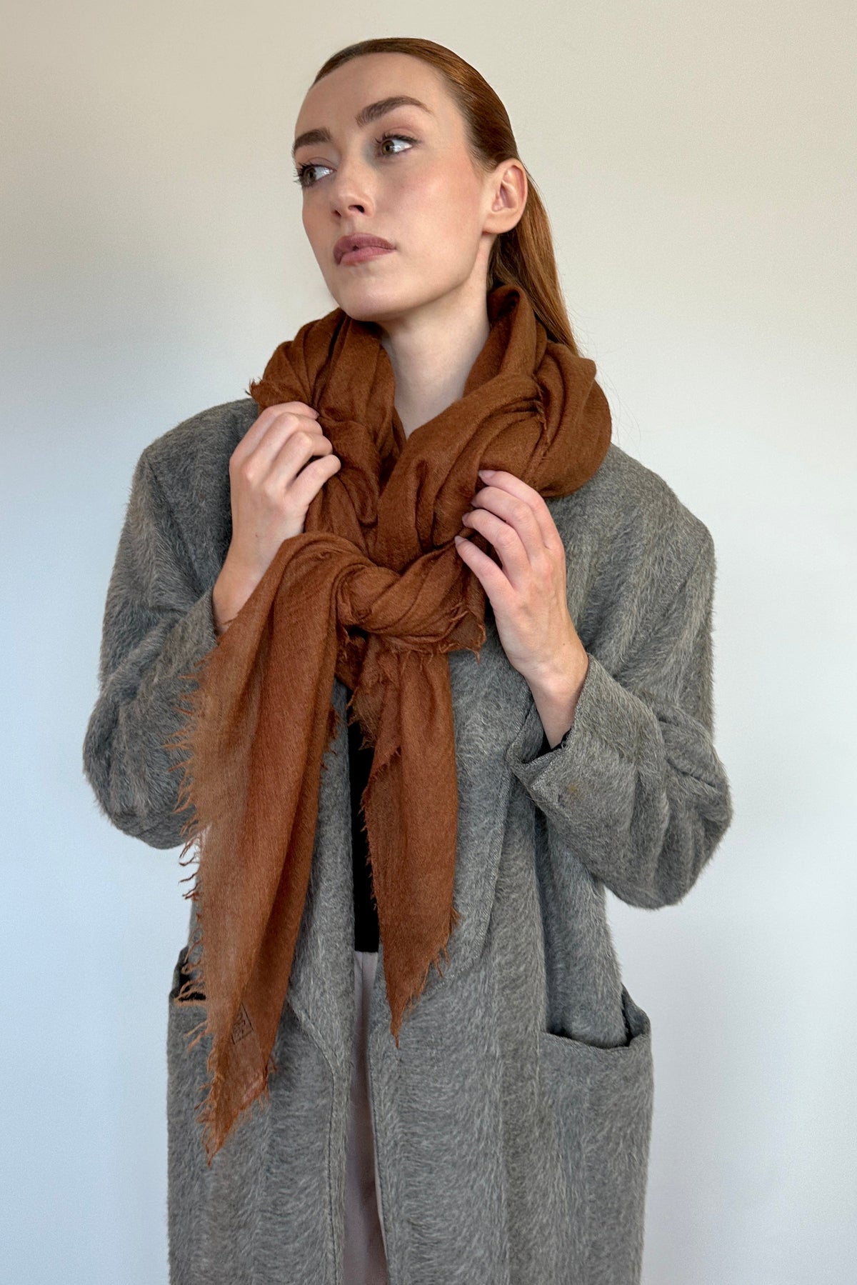 A woman wearing a brown cashmere scarf wrapped around her neck and a gray coat, posing against a plain light gray background.