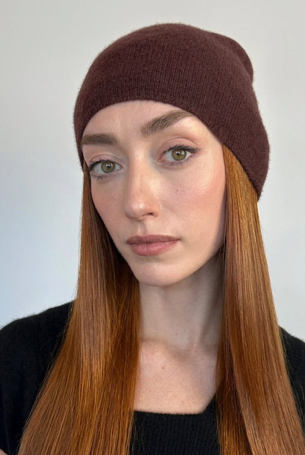 A woman with long red hair wearing a dark brown knit beanie and a black top, photographed against a plain light gray wall.