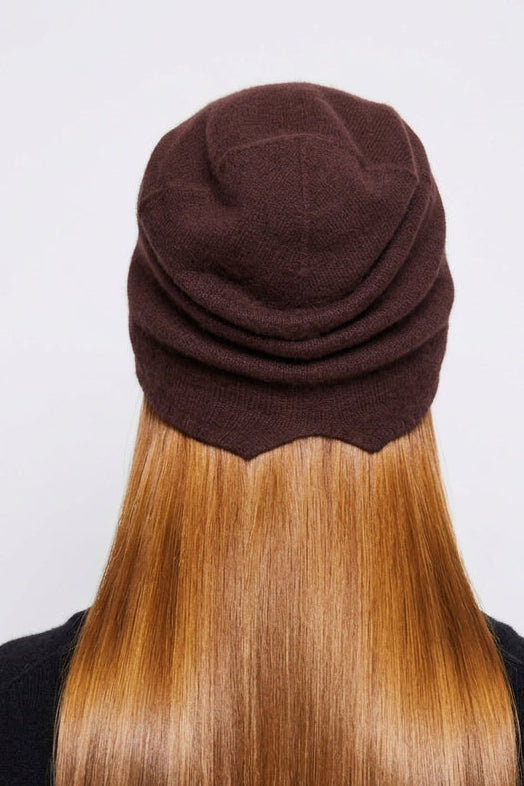 A person viewed from behind wearing a dark brown knitted beanie hat, with long light brown hair and a black top against a plain light gray background.