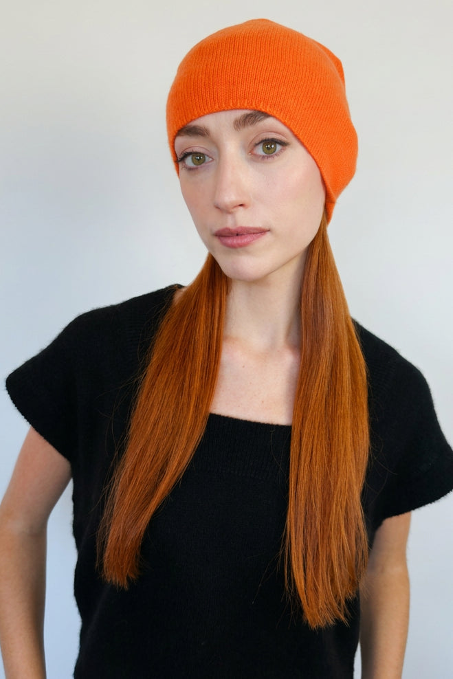 A woman with long auburn  hair wearing a bright orange cashmere knit beanie and a black short-sleeve top, standing against a plain light gray wall.
