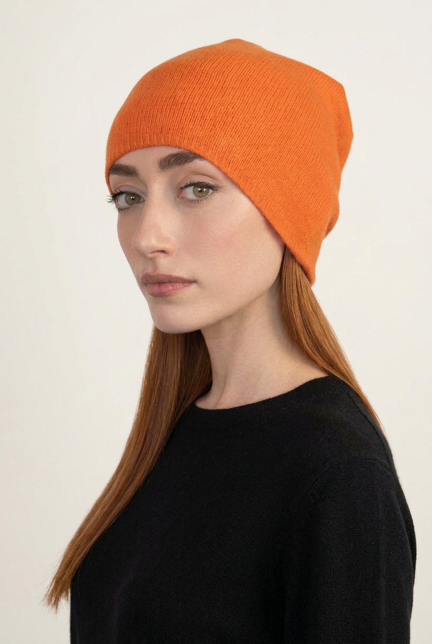 A woman with long red hair wearing a bright orange knit beanie and a black short-sleeve top, photographed against a plain light gray wall.