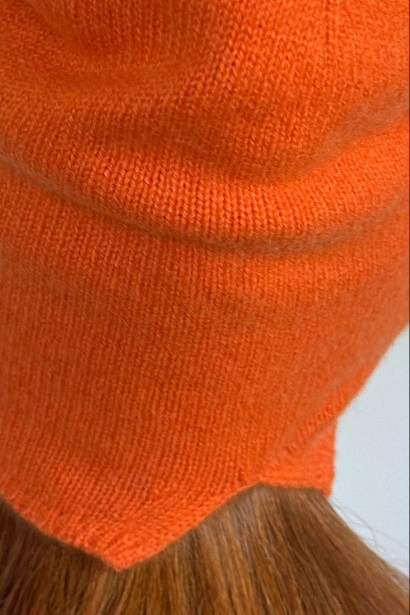 An orange knit beanie hat worn by a person, viewed from the back, with long light brown hair visible below the hat against a plain light gray background.