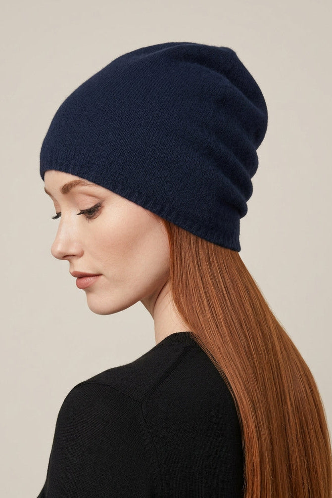 A woman with long red hair is shown in profile wearing a dark navy blue knit beanie and a black top against a plain light beige studio background.