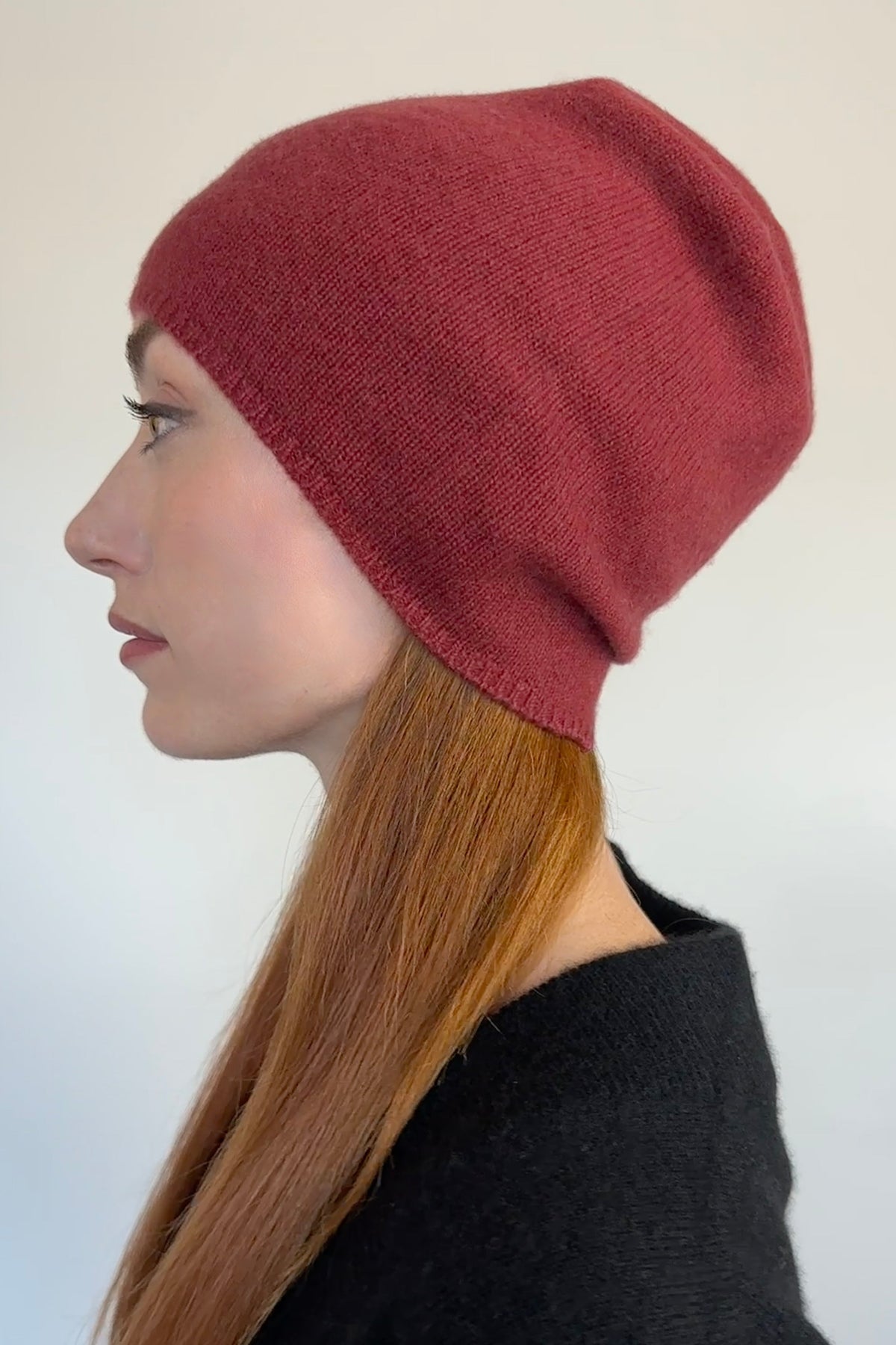 A woman in profile wearing a ribbed knit beanie hat in a muted brick red color, paired with a black top against a plain light background.