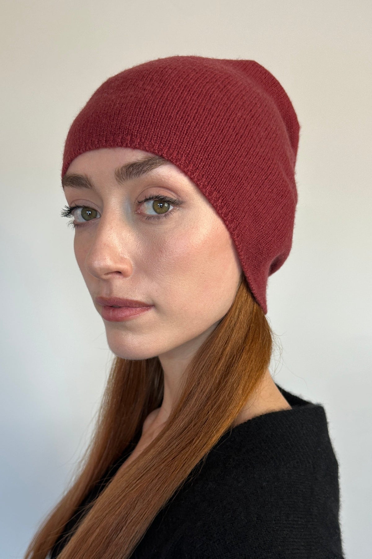 A woman with long auburn hair wearing a ribbed knit beanie in a deep burgundy red, paired with a black knit top, photographed against a neutral light gray background.