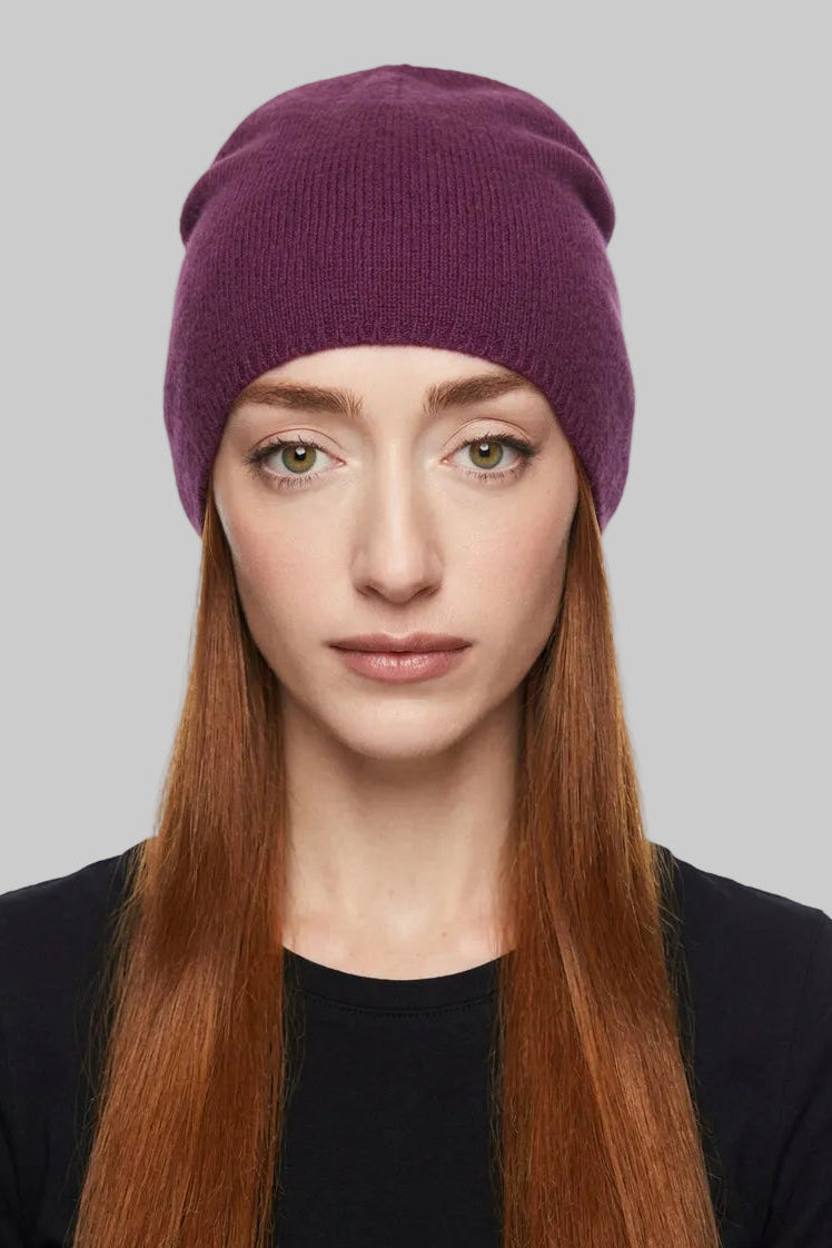 A woman with long light brown hair wearing a slouchy knit beanie in a deep plum purple color, paired with a black top, photographed against a plain off-white background.