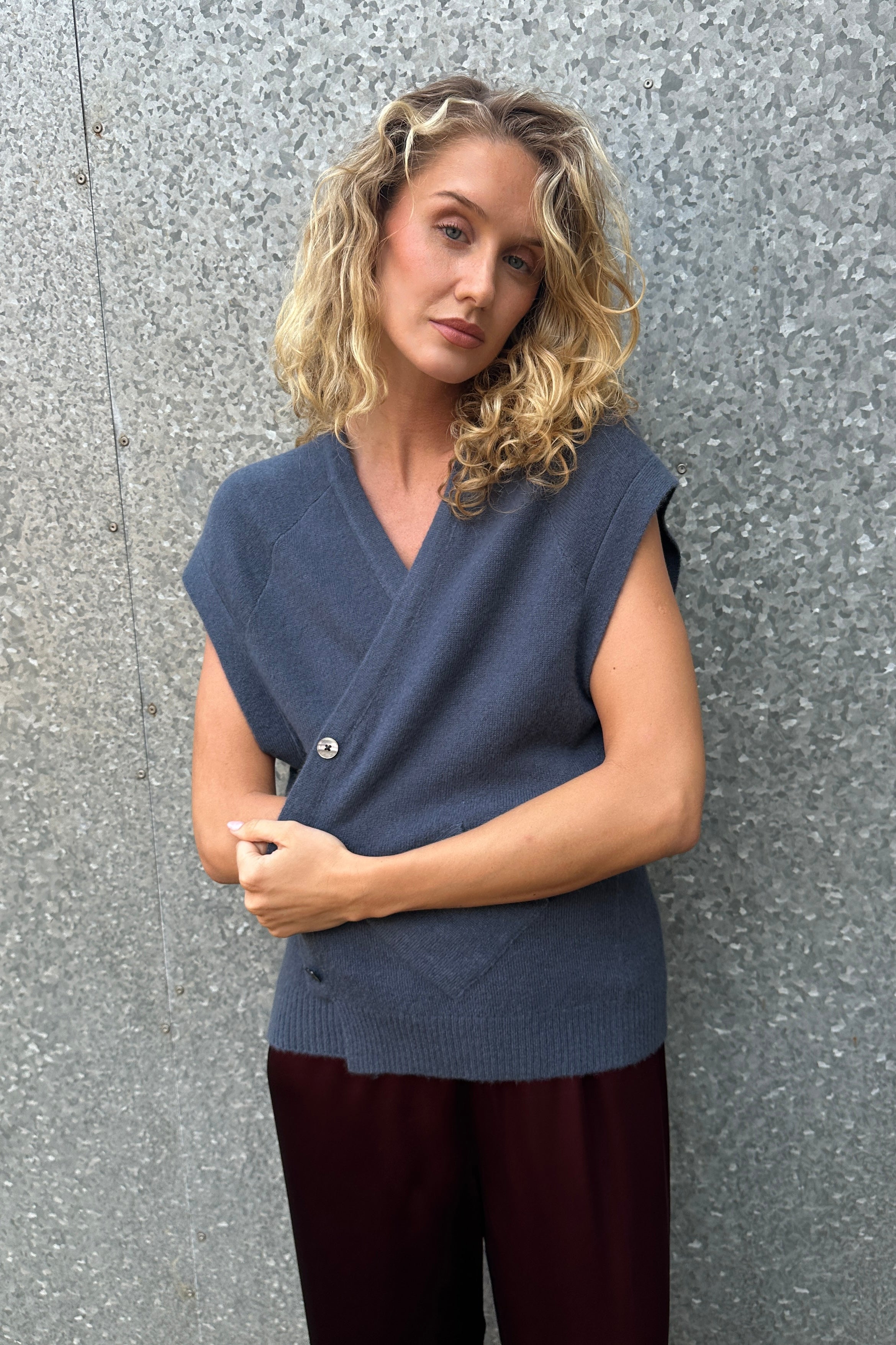 model wearing daisy blue  cashmere sleeveless cardigan  and plaid scarf against a textured wall.