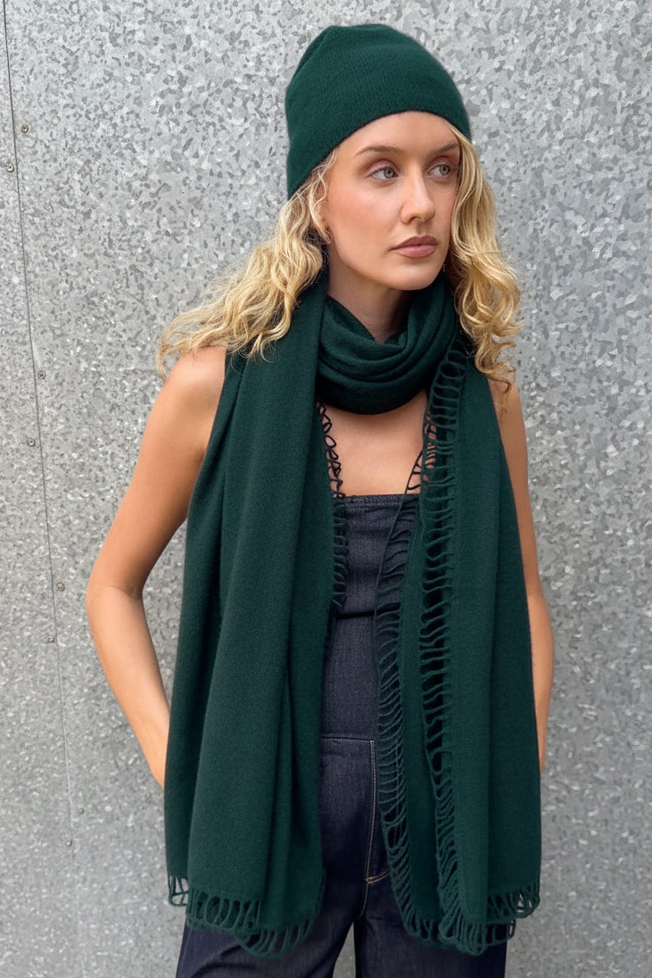 Woman wearing a dark green scarf and beanie against a gray wall