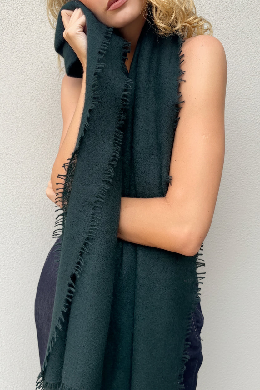A woman wearing a dark forest green fringed scarf draped over her shoulders, standing against a textured gray concrete wall.