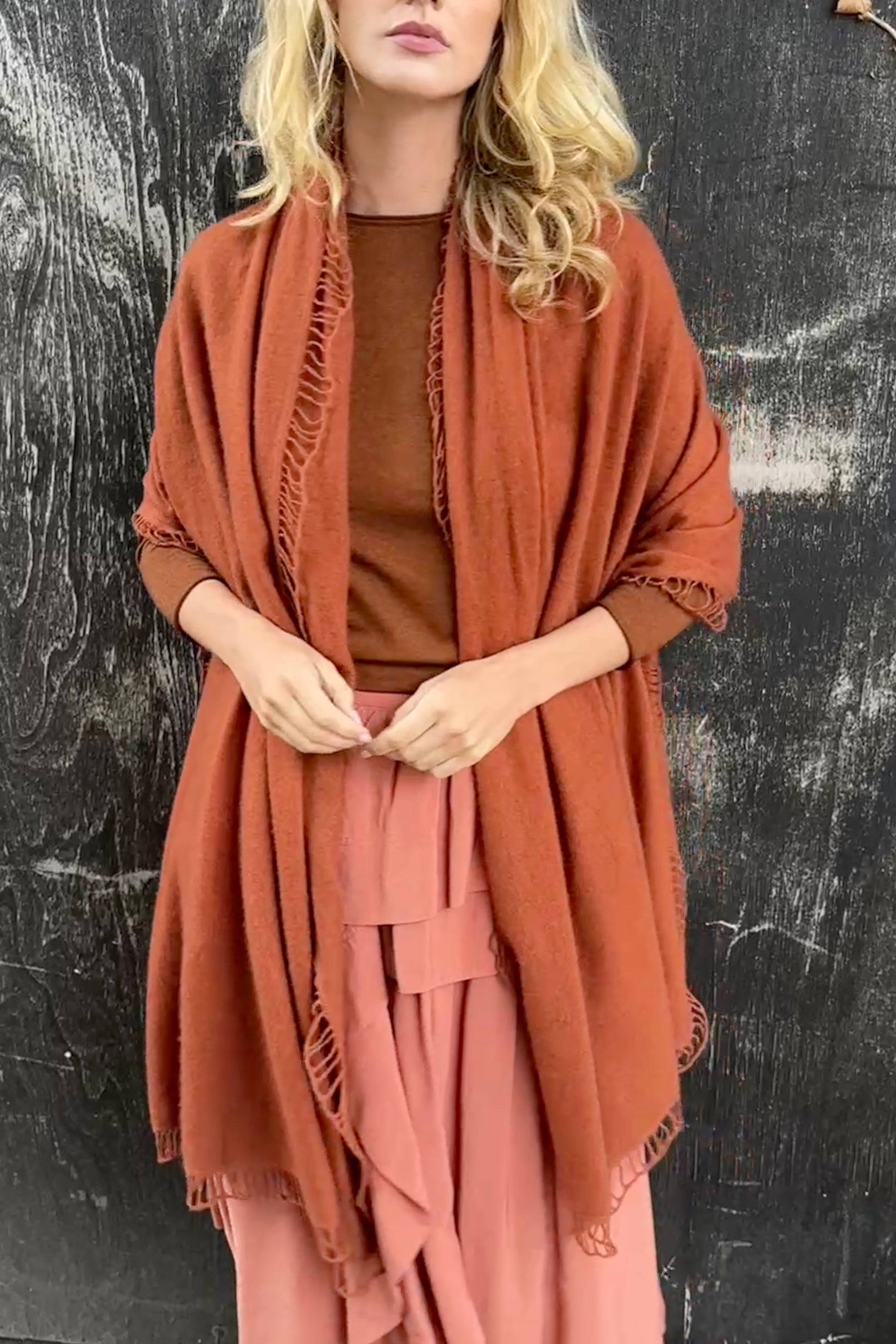 Woman wearing a rust-colored shawl over a brown top and pink pants against a black textured background