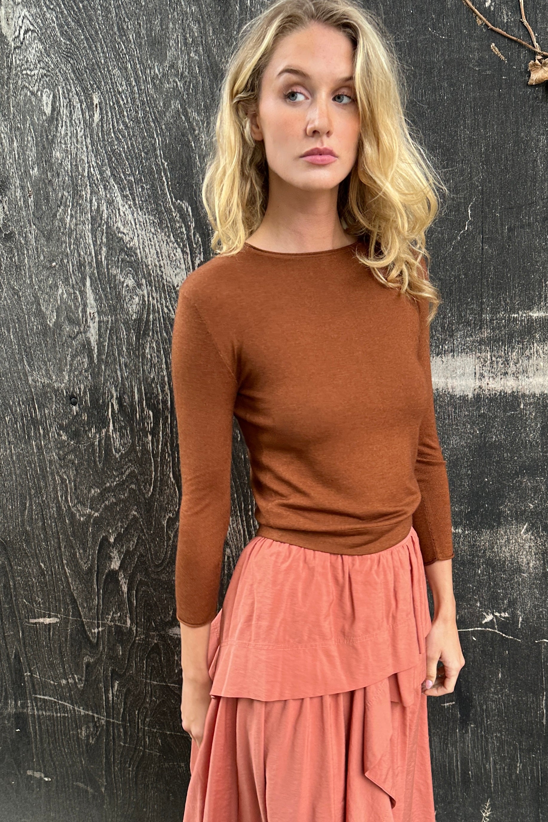 model wearing a rust  brown  cashmere silk top or sweater and pink skirt against a textured black wall