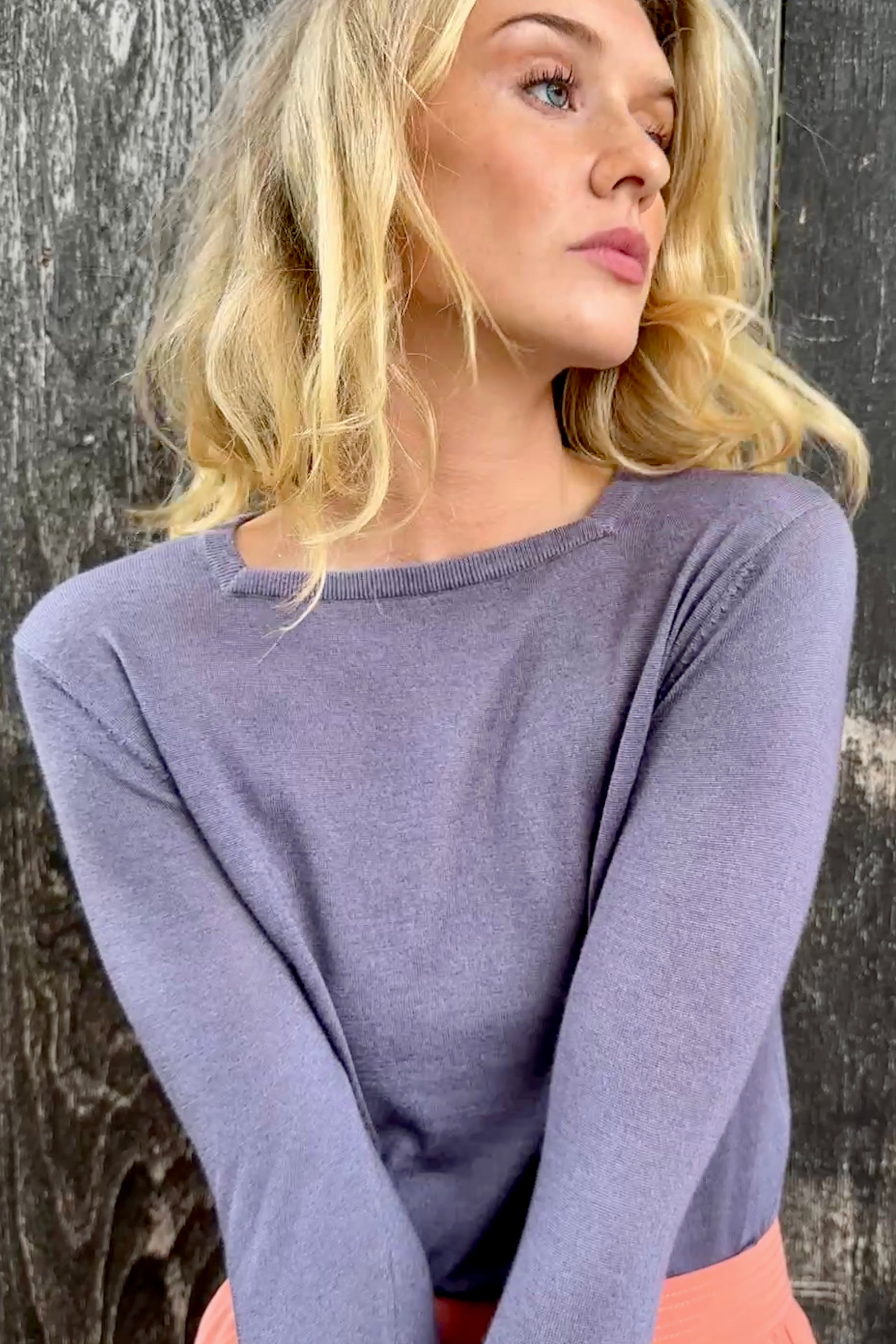 Model wearing a purple  lassa square neck long-sleeve top and pink skirt against a textured black background close up