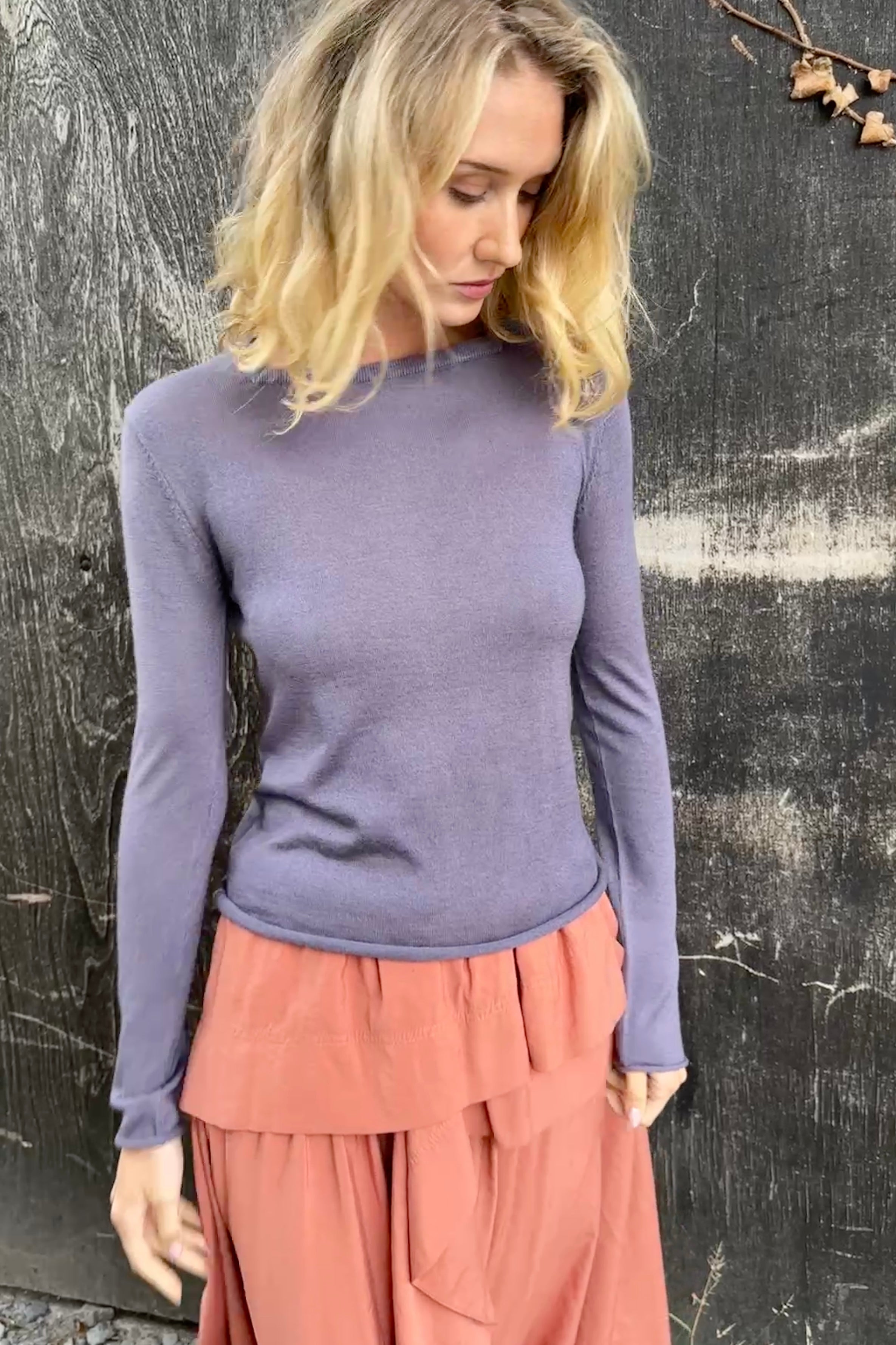 Model wearing a purple  lassa square neck long-sleeve top and pink skirt against a textured black background side view