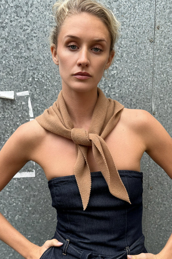 Woman wearing a strapless dark blue top and high-waisted pants with a beige scarf around her neck, standing against a gray wall.