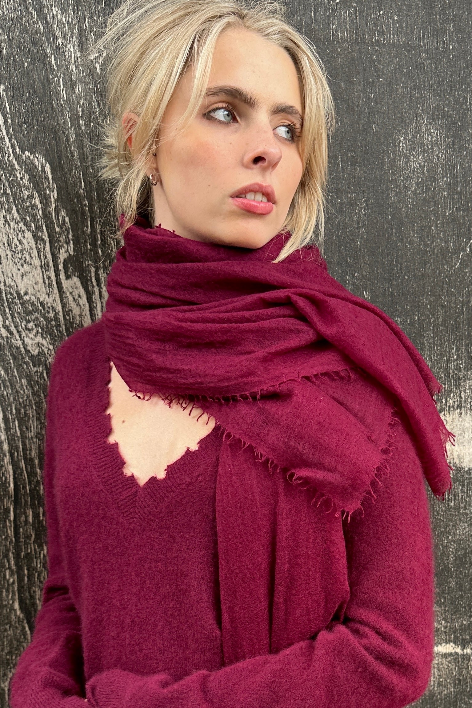 Woman wearing a burgundy  cashmere marmee scarf and sweater against a textured wall.