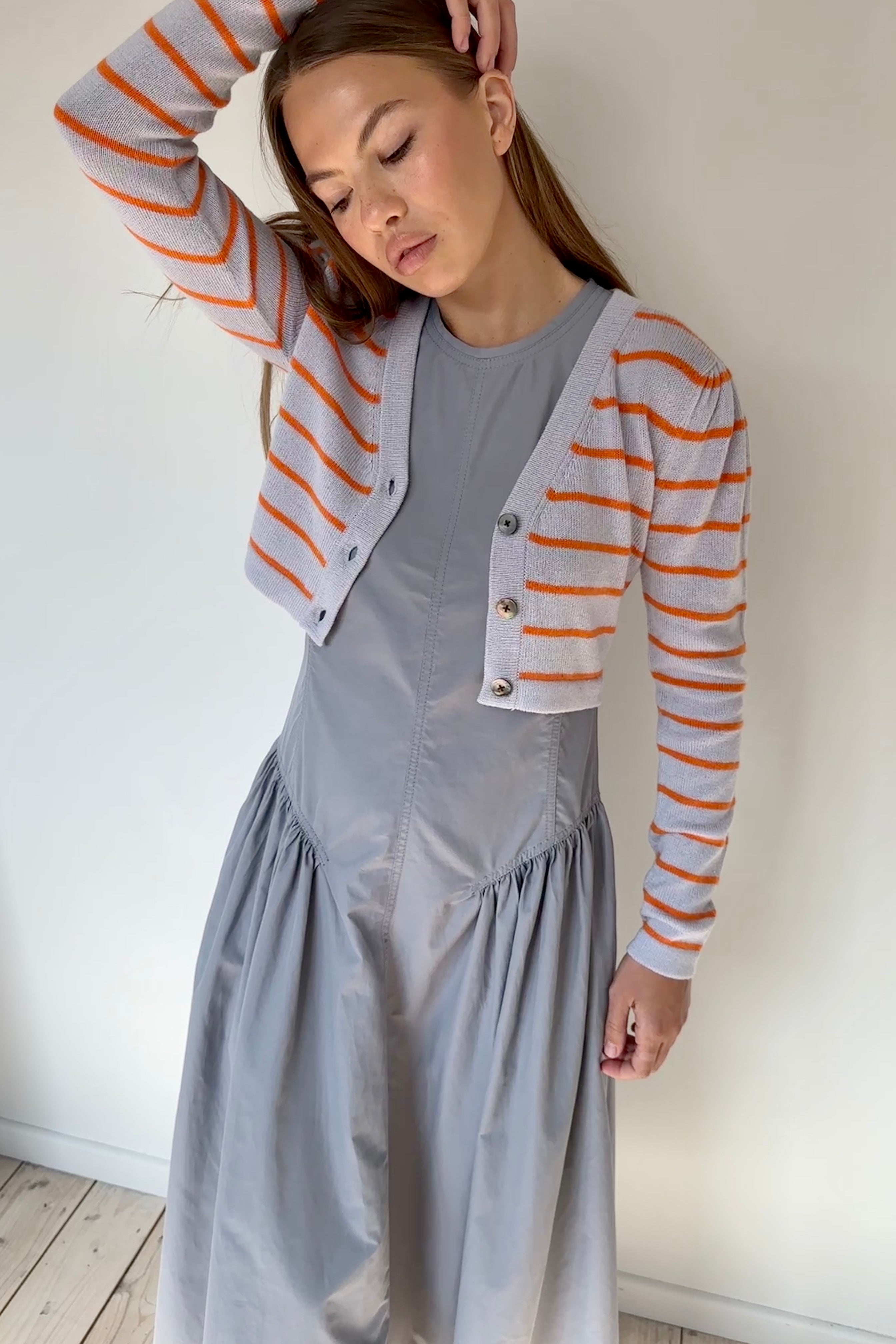 Woman wearing a gray dress with an orange and white striped cardigan against a white wall.