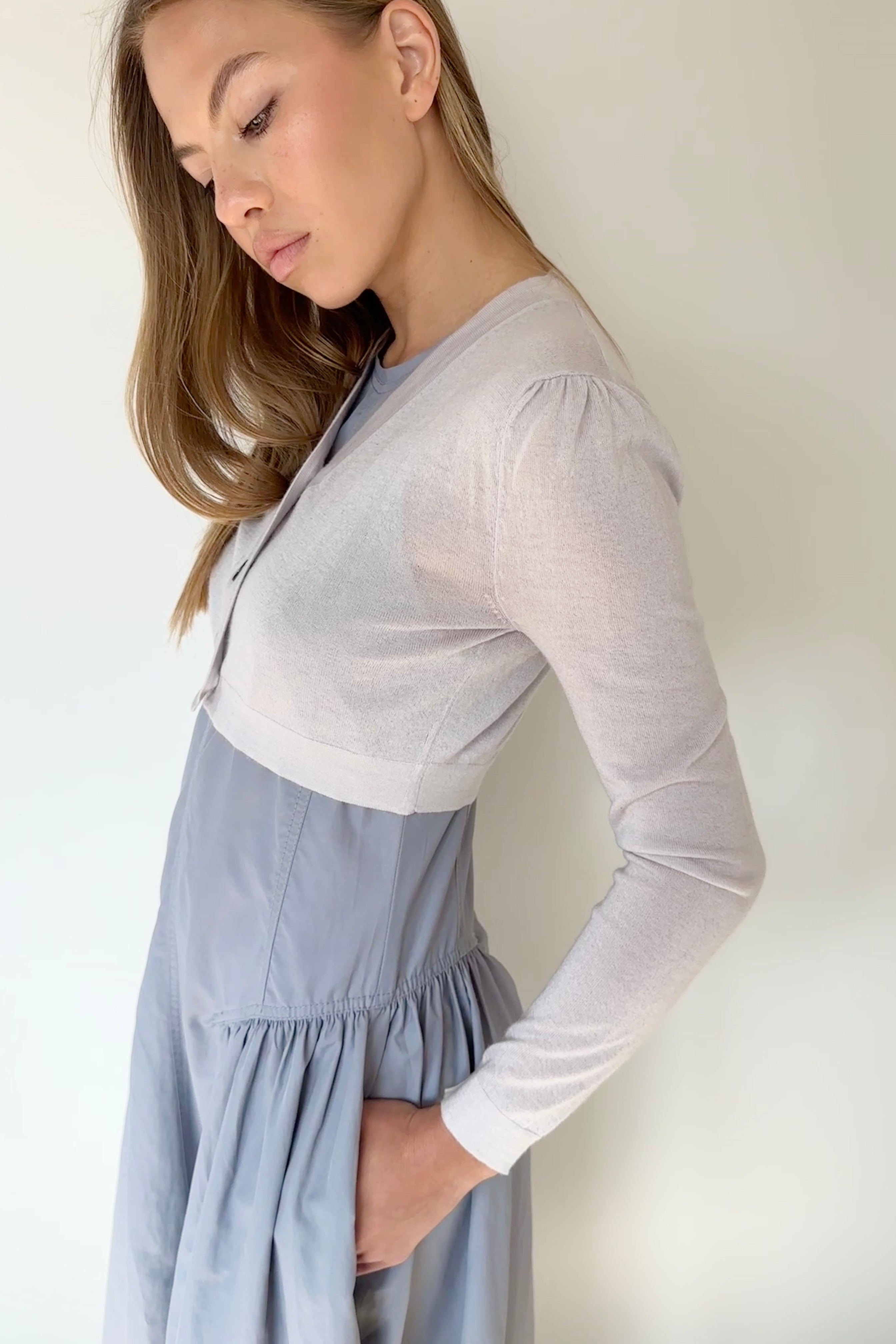 Woman wearing a light gray long-sleeve top over a blue dress against a plain background