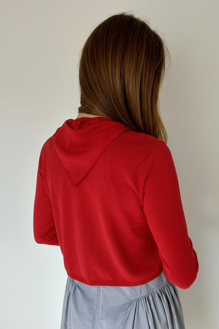 A woman viewed from behind wearing a red cardi and a dusty blue skirt, standing against a plain off-white wall.