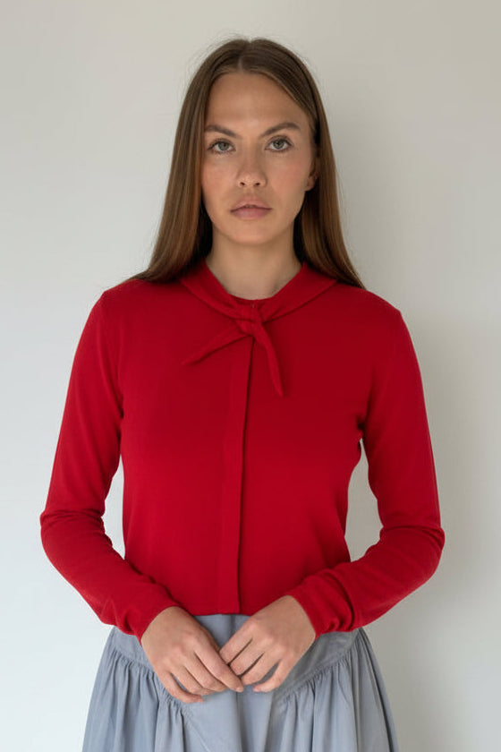A woman modeling a redwith a tie-neck detail, paired with a dusty blue skirt, standing against a plain off-white wall.