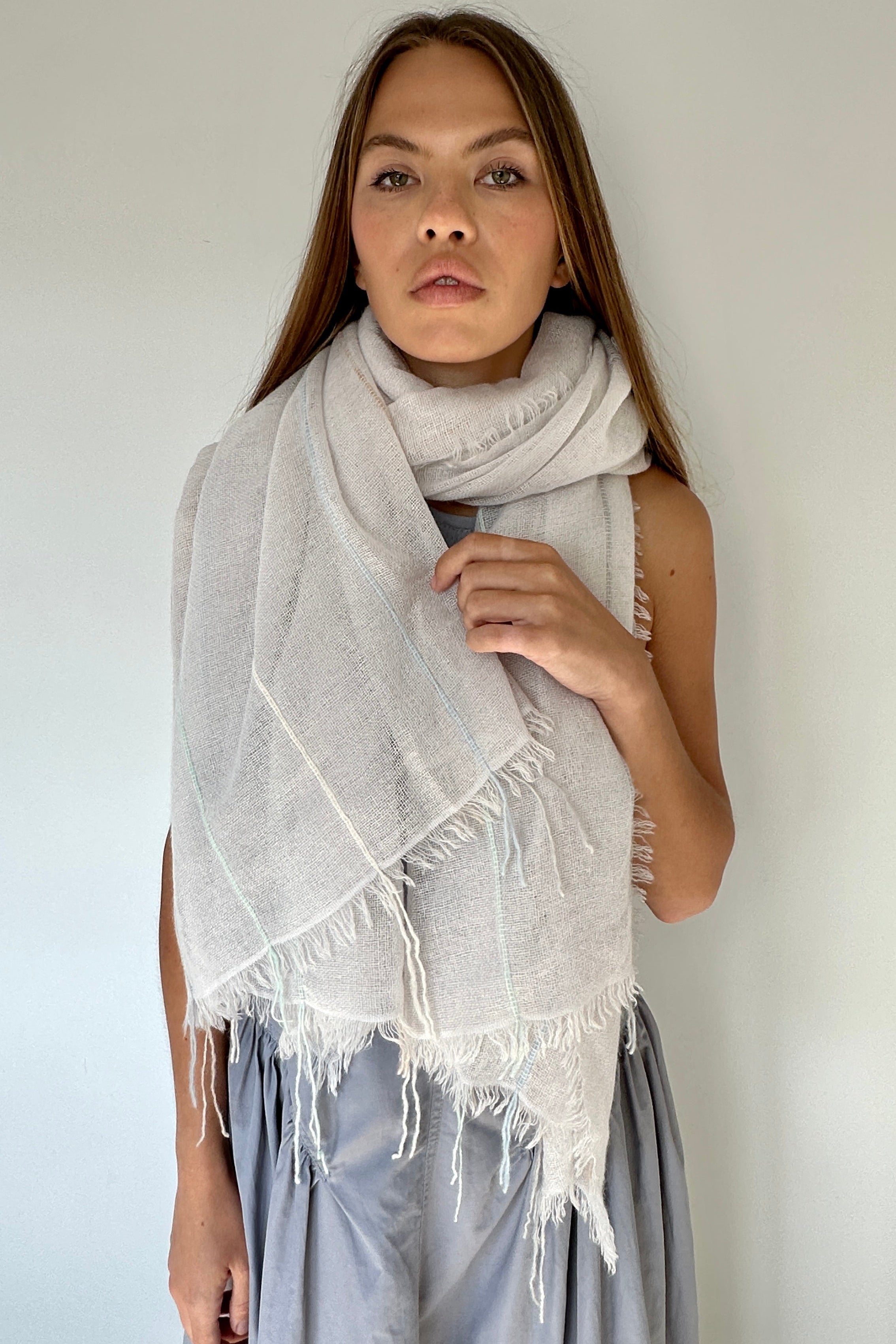 Woman wearing a light gray scarf with white fringe against a plain background