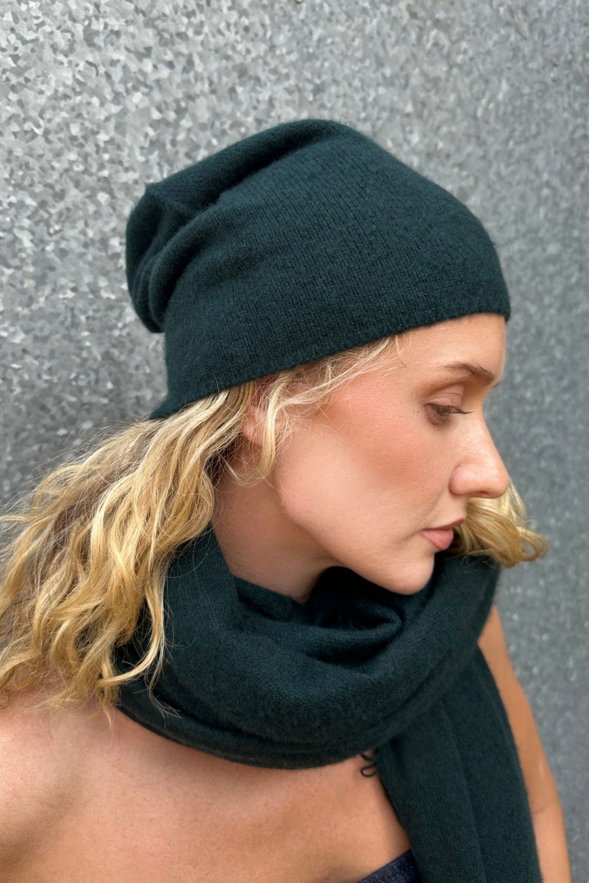 model wearing dark teal bottle greeen blue beanie and scarf against a gray background