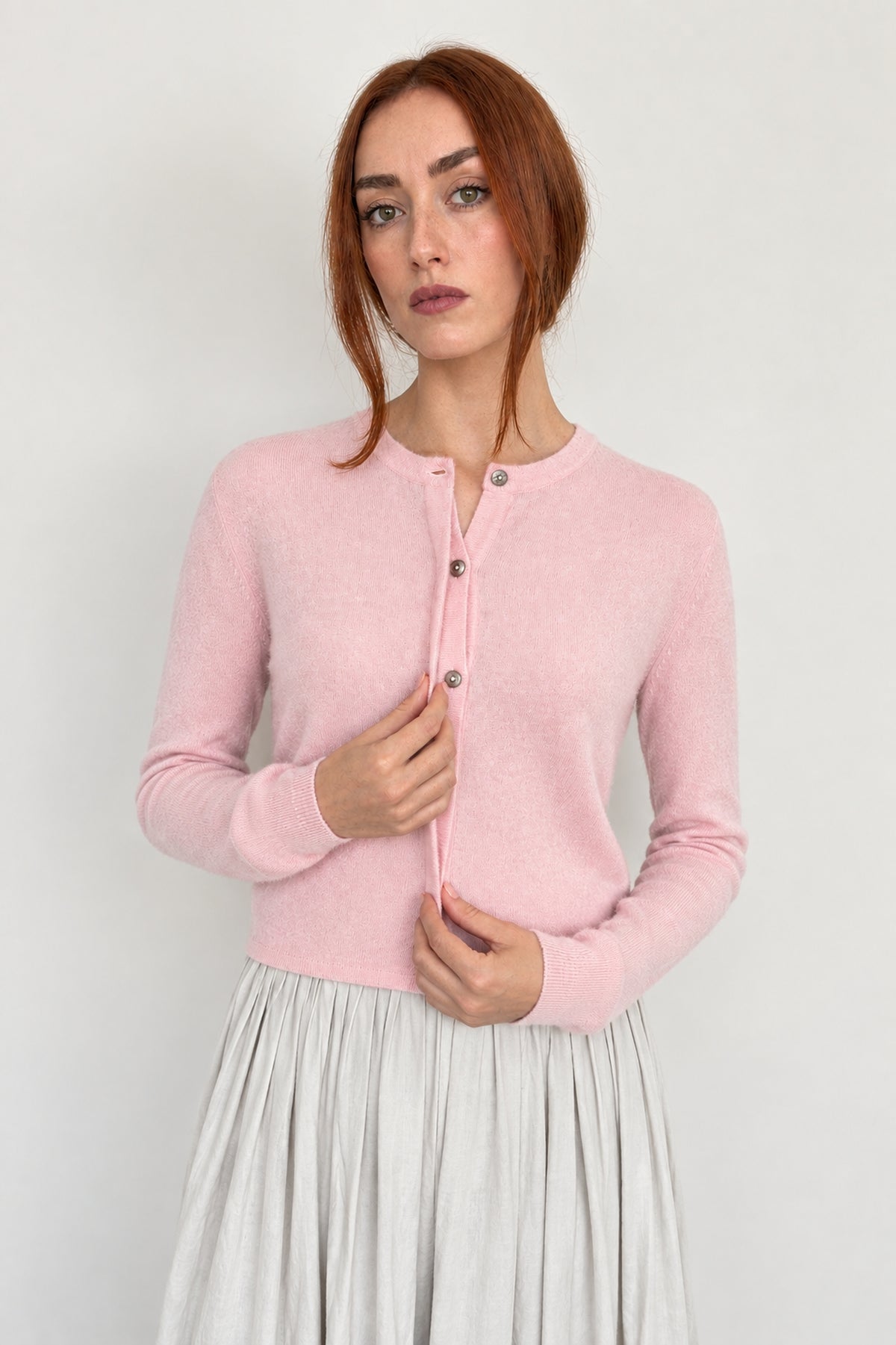 A woman with red hair wearing a light pink long-sleeve cardigan with a buttoned stand collar and a light gray pleated skirt, standing against a plain light gray background.