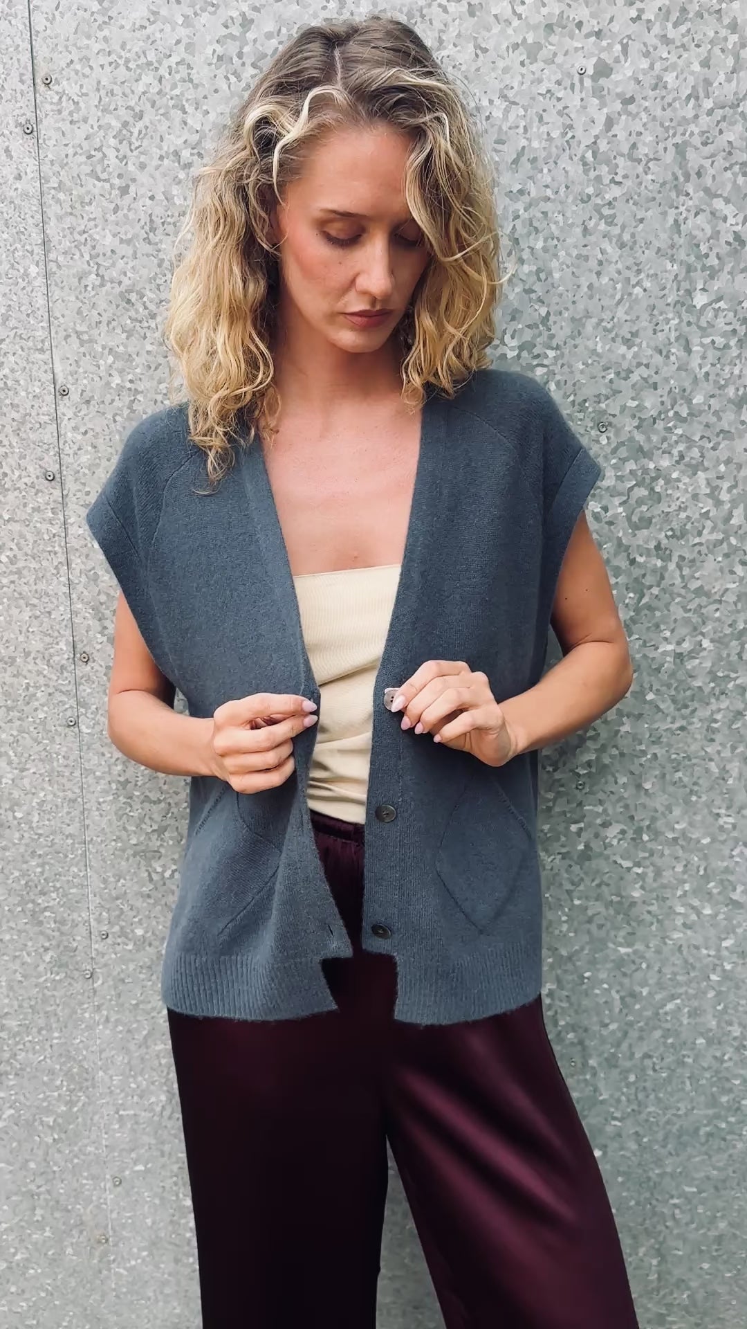 model wearing  daisy blue  cashmere sleeveless cardigan  and plaid scarf against a textured wall.